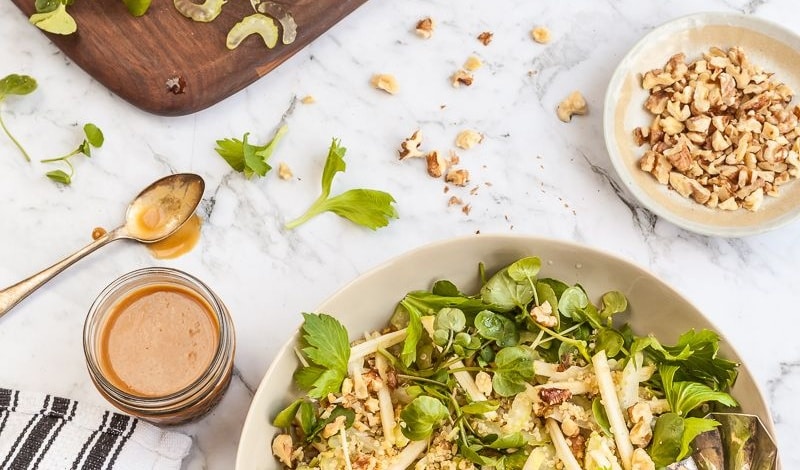 Chopped celery, watercress, toasted walnuts, and honey miso dressing laid out next to a bowl of celery apple walnut salad