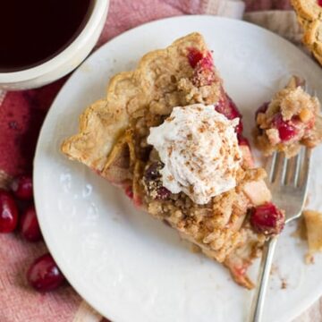 Overhead view of a slice of Chai-Spiced Cranberry Apple Pie topped with whipped cream on a white plate, with coffee and fresh cranberries alongside