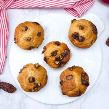 Five date and apple muffins on a round white marble board, surrounded by chopped dates and fresh apples