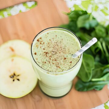 Overhead close-up of a green apple smoothie in a glass with a straw, garnished with cinnamon, next to sliced Granny Smith apple and fresh spinach