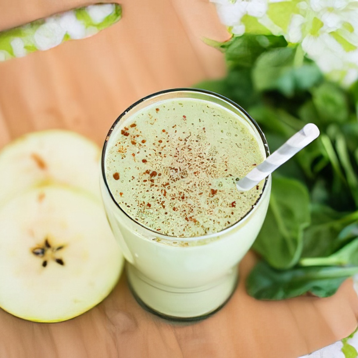 Overhead close-up of a green apple smoothie in a glass with a straw, garnished with cinnamon, next to sliced Granny Smith apple and fresh spinach