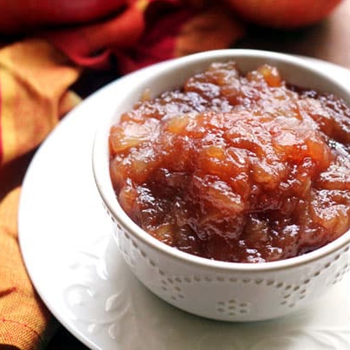 A square, close-up view of a white bowl of thick, deep brown homemade apple butter with whole apples in the background