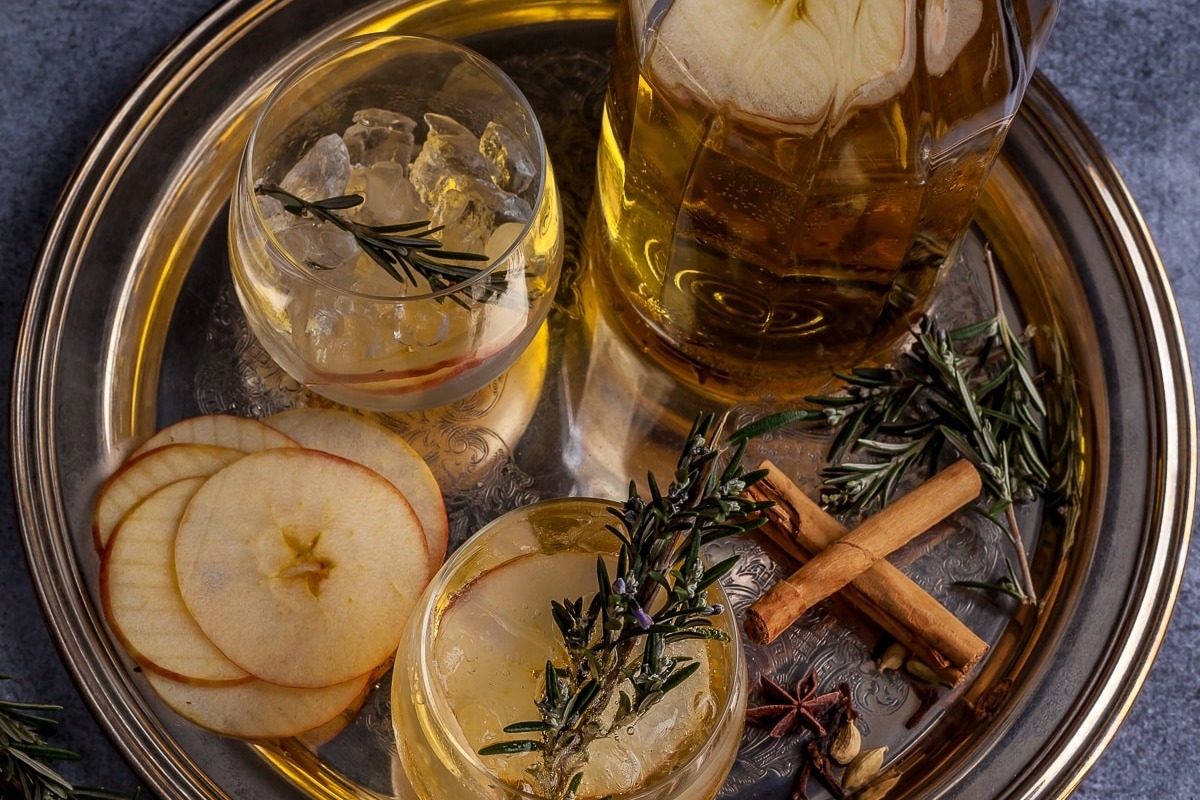 Overhead view of sparkling mulled apple cider in glasses on a silver tray with apple slices, cinnamon sticks, star anise, and rosemary