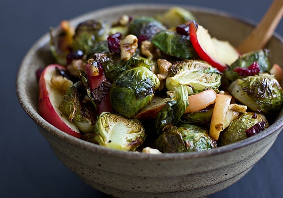 Bowl of roasted brussels sprouts and apples with walnuts, cranberries, and a wooden spoon