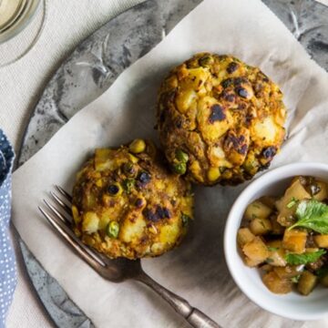 Overhead view of two golden-brown samosa cakes on parchment with a bowl of apple chutney