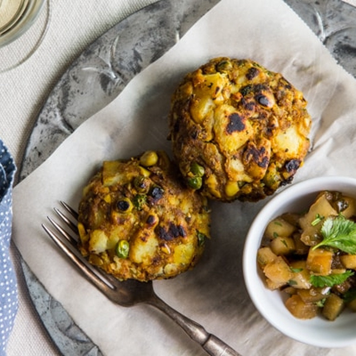 Overhead view of two golden-brown samosa cakes on parchment with a bowl of apple chutney