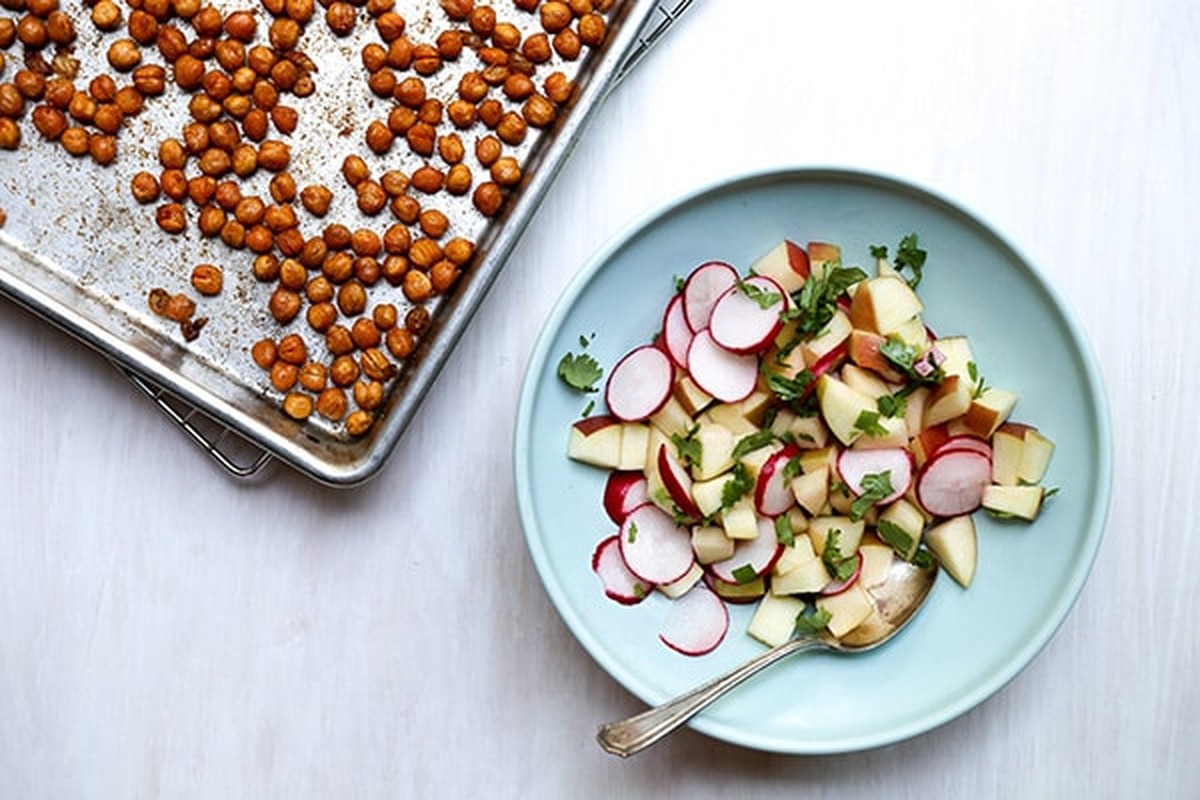 A sheet pan of roasted chickpeas next to a blue plate piled with apple-radish slaw and cilantro