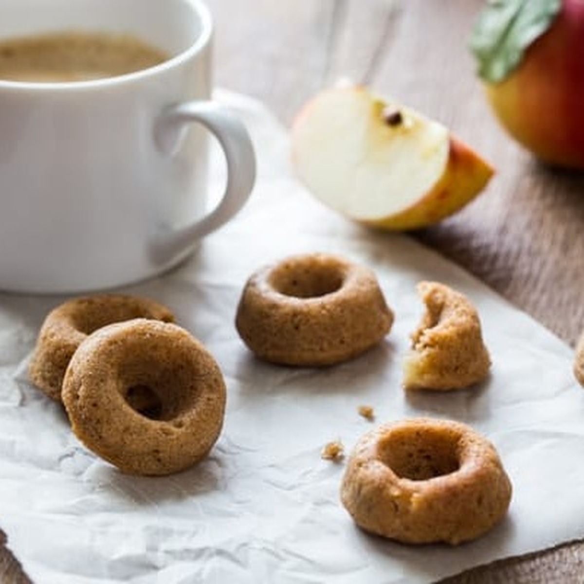 Vegan apple cider doughnuts on parchment paper with coffee and a sliced apple
