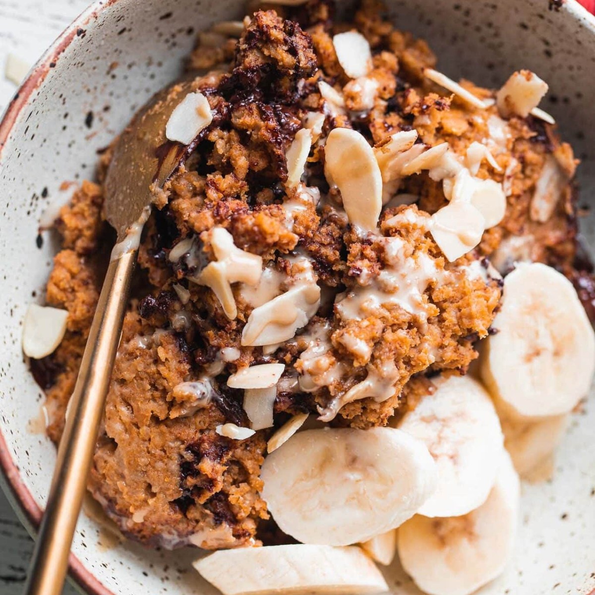 Bowl of vegan pumpkin baked oatmeal with banana slices, sliced almonds and a gold spoon