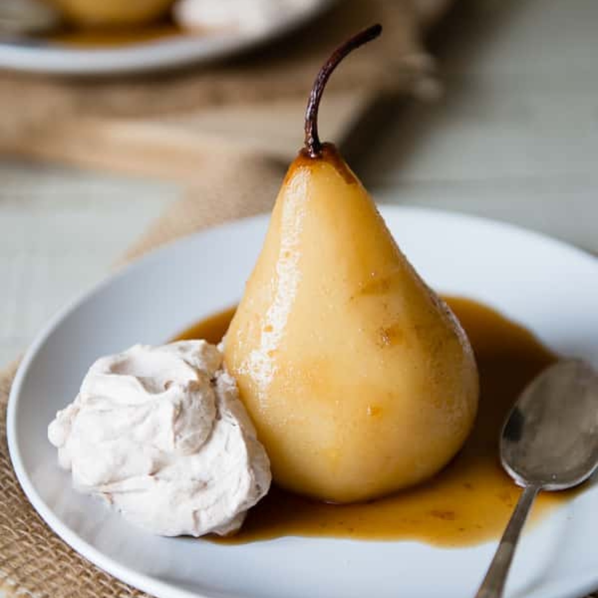 Chai poached pear standing in amber syrup on a white plate with a swirl of cinnamon whipped cream and a spoon