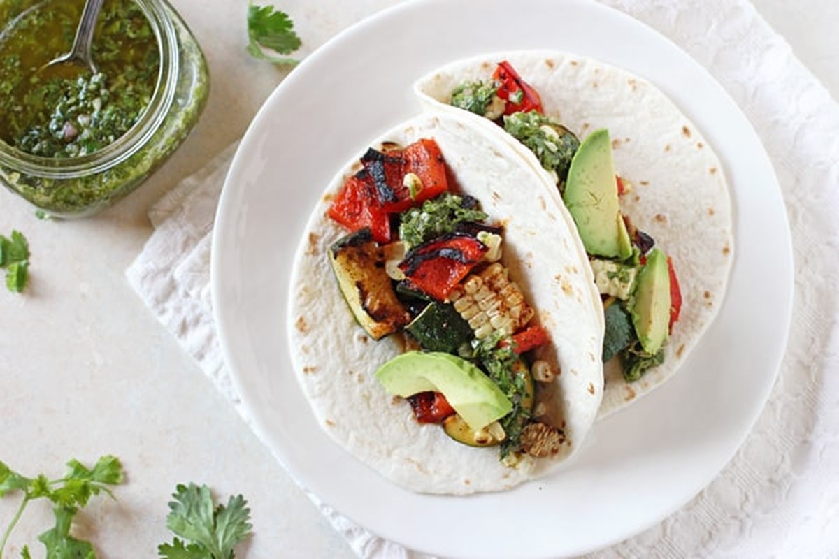 Two grilled veggie tacos on a white plate next to a jar of chimichurri sauce