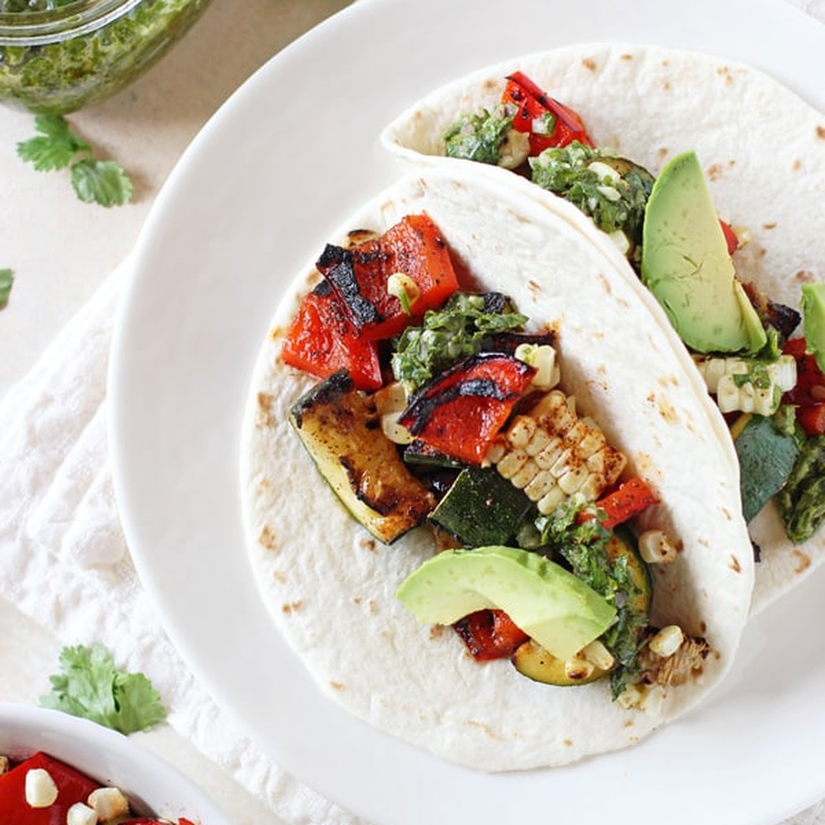 Two grilled veggie tacos on a white plate filled with charred red pepper, zucchini, corn and avocado, drizzled with chimichurri