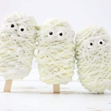 Three Halloween mummy cakesicles decorated with white chocolate drizzle and candy eyes