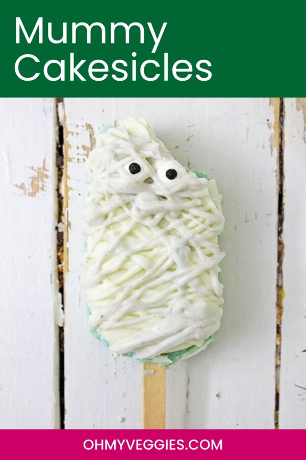 Mummy cakesicle with candy eyes on a rustic white board, with text overlay for Pinterest