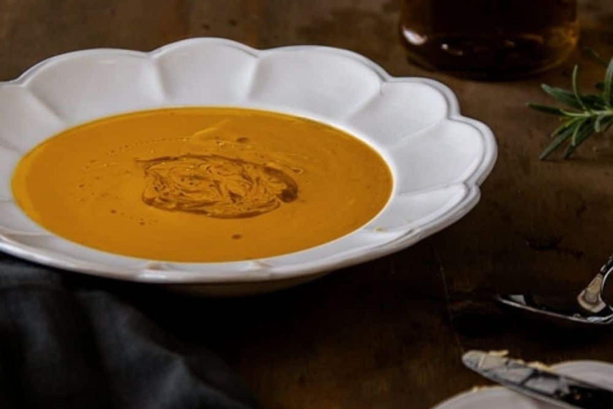Bowl of roasted butternut pumpkin soup with a swirl of olive oil, fresh rosemary and toast on a wooden table