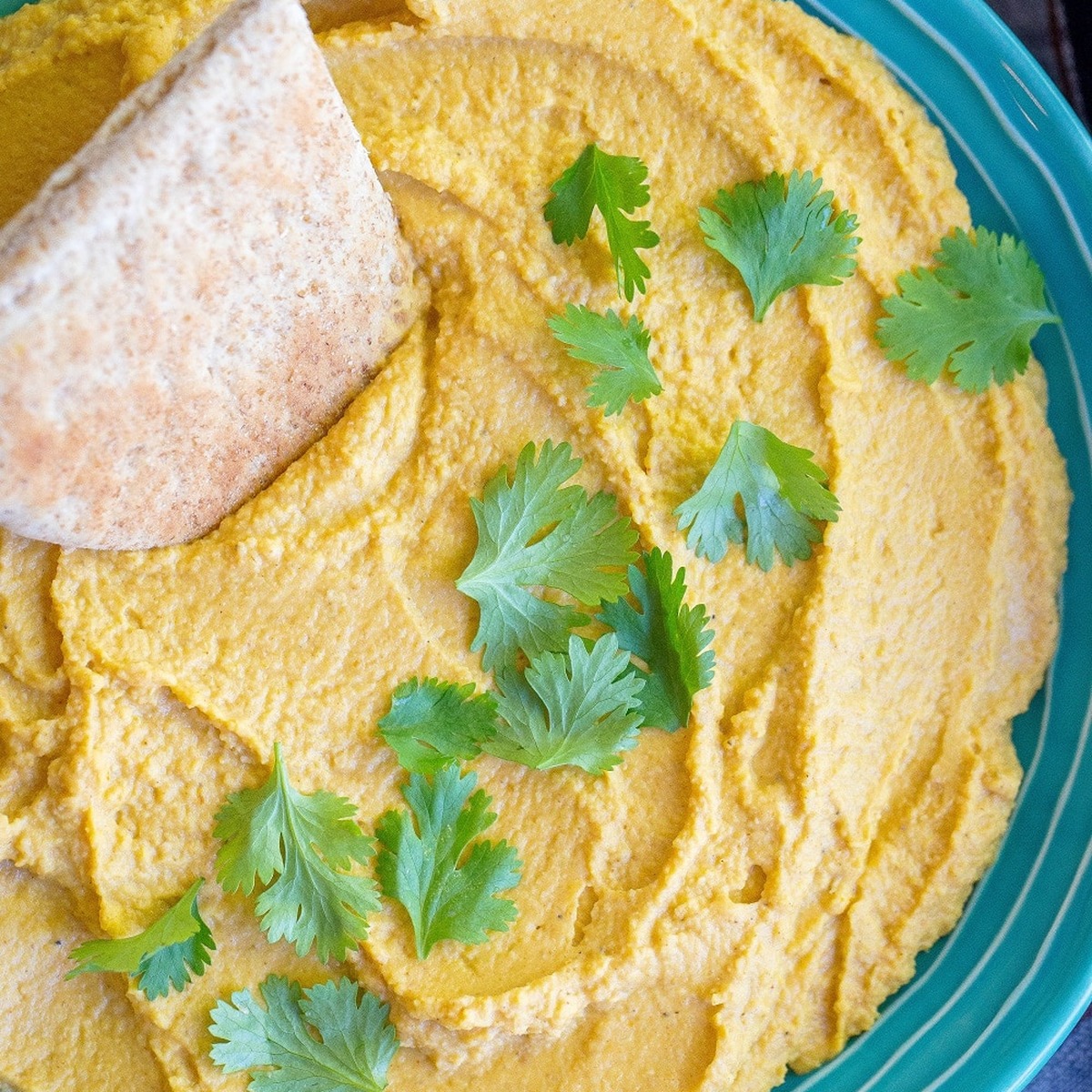 Pumpkin hummus swirled in a teal bowl with fresh cilantro leaves and a pita wedge