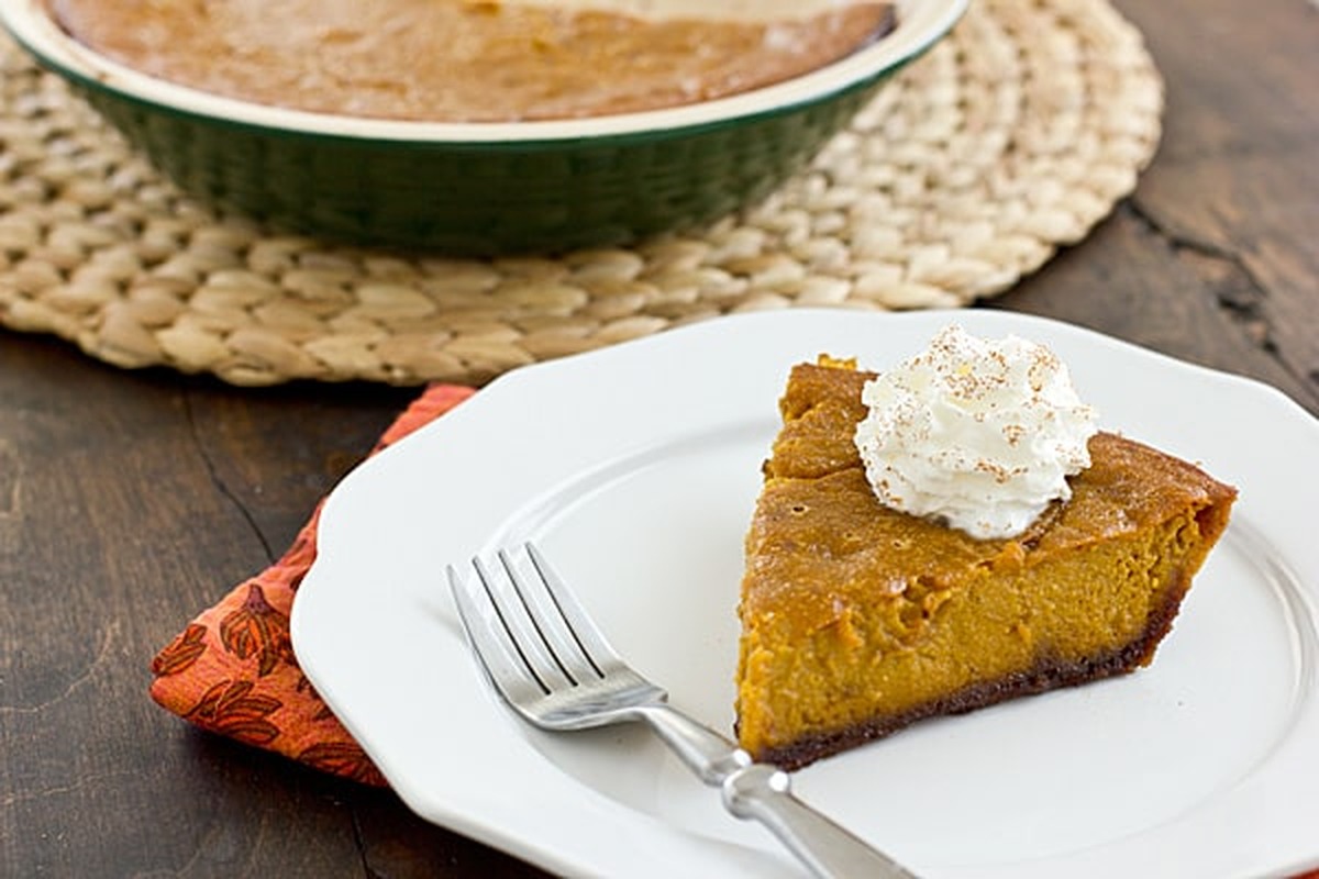 Slice of pumpkin pie with a gingersnap crust and whipped cream on a white plate, with the whole pie in a green dish behind it
