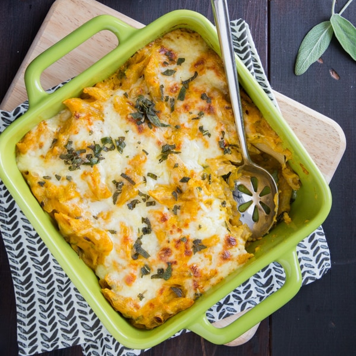 Overhead view of pumpkin sage baked ziti in a green square baking dish topped with melted white cheddar and crispy sage